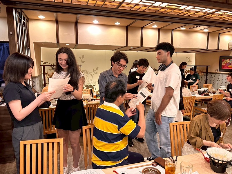 Students and local residents smile and interact while playing a paper-based game in a traditional Japanese restaurant