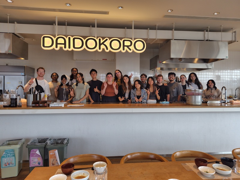 A group of CET Japan students smiling in front of a sign that says "DAIDOKORO" for a cooking class