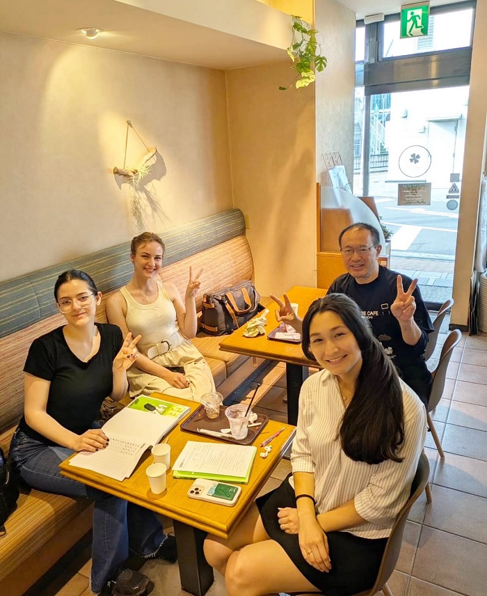 Four students sit around a table at a cozy café, smiling and holding up peace signs with study materials in front of them