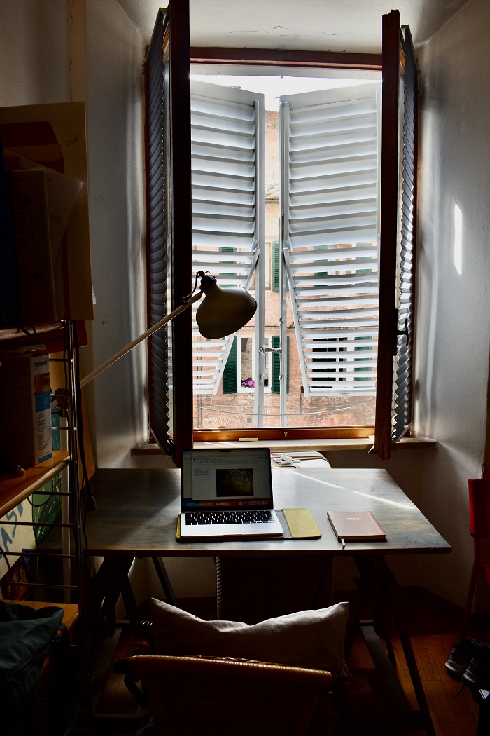 A desk with a laptop opened and notebooks and open windows in a bedroom