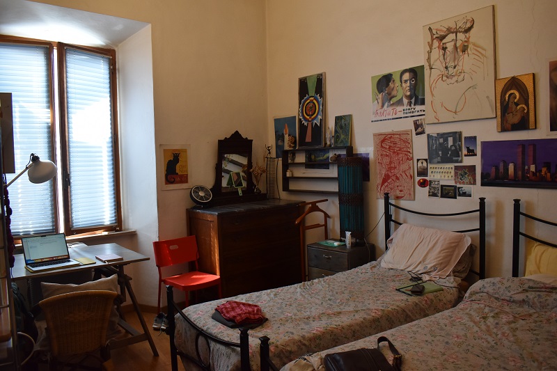 Two beds, a desk and chair, a dresser, a wardrobe, and big window with shades down in a homestay room at CET Siena