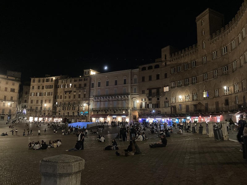 People n standing or sitting in the Piazza del Campo with friends, drinking some wine or beer or just chatting at night