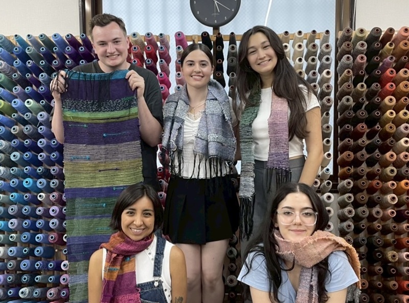 A group of CET Japan students wearing their creations from a saori weaving workshop while standing in front of a wall of different colored threads