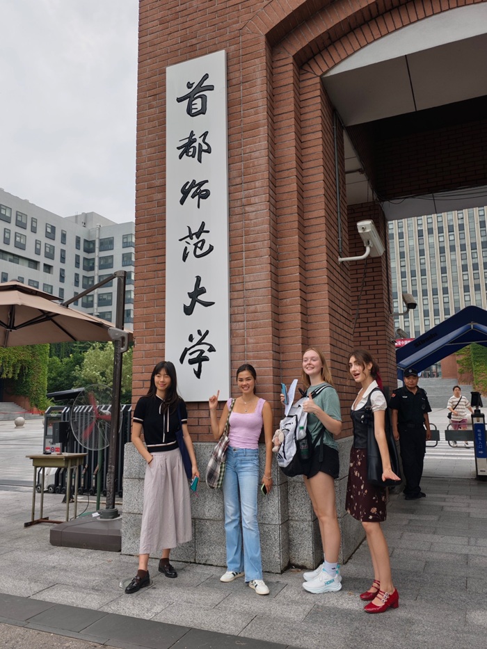 Four CET Beijing students smiling and posing by a pillar of a building at Capital Normal University with Chinese characters written on the pillar