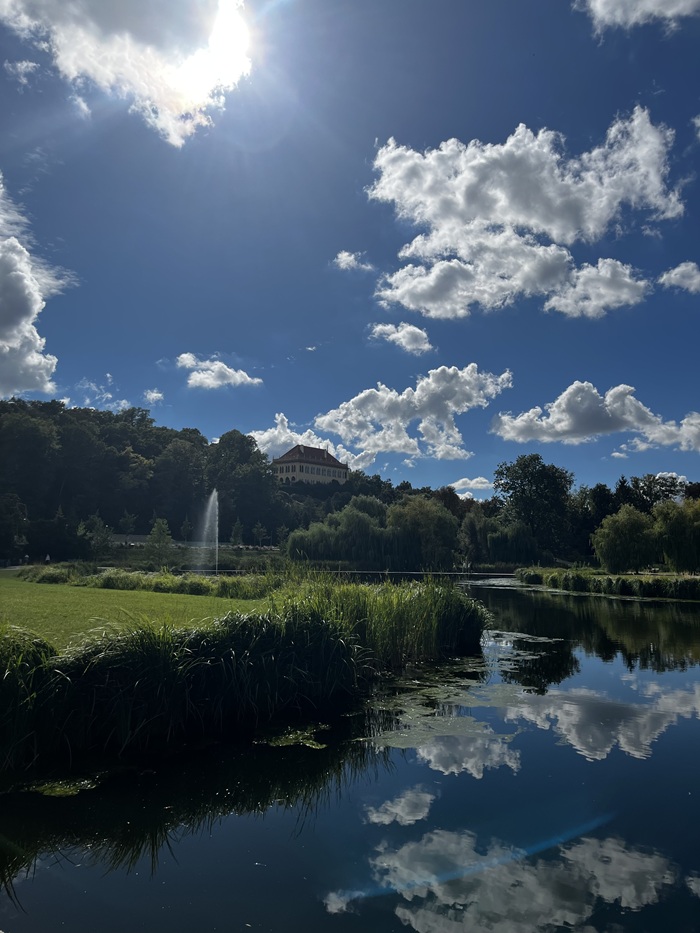 An empty park scene in Prague with water, grass, and fountain under a partly cloudy day