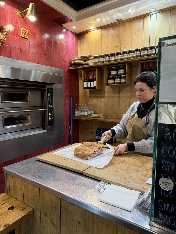 A shop-worker, surrounded by ovens, wines, and jars, about to cut up a sandwich in a store