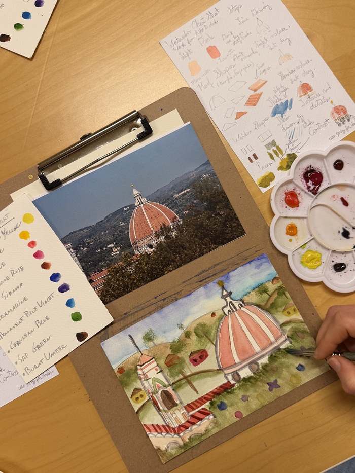 A table displaying a photo of the Duomo in Florence above a painting of the same scene, alongside a sheet of sketches and a paint palette