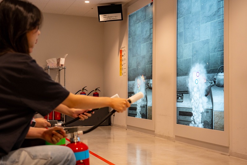 CET Japan students using simulated fire extinguishers to extinguish virtual fires displayed on large screens at the Osaka City Abeno Disaster Prevention Center.