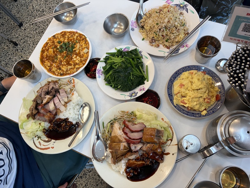 A table with a variety of foods on plates including BBQ pork and roasted duck rice, Mapo tofu, Stir-fried leafy greens, and fried rice.