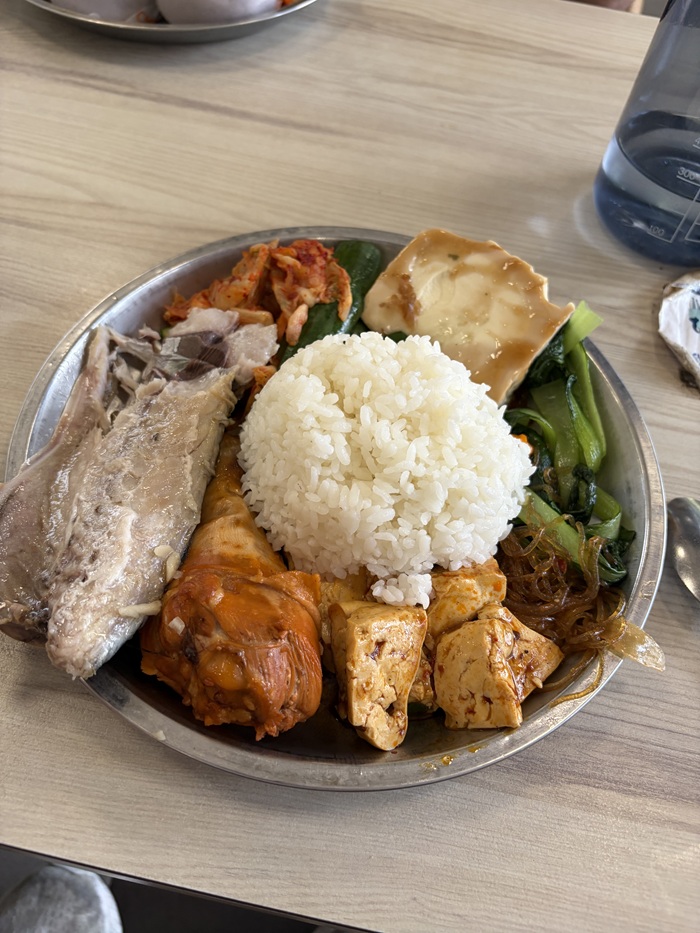 This plate features a central scoop of white rice surrounded by various side dishes like steamed fish, braised chicken drumstick, tofu, and more.