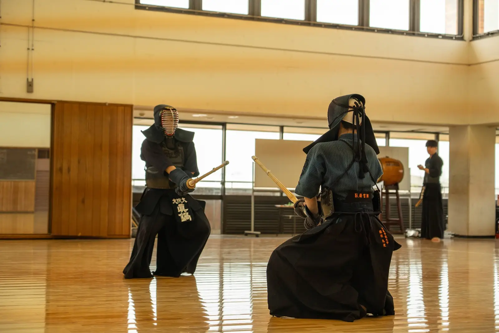 Two people in uniform in a kendo session holding shinai (bamboo swords) inside of a wooden-floored gymnasium.