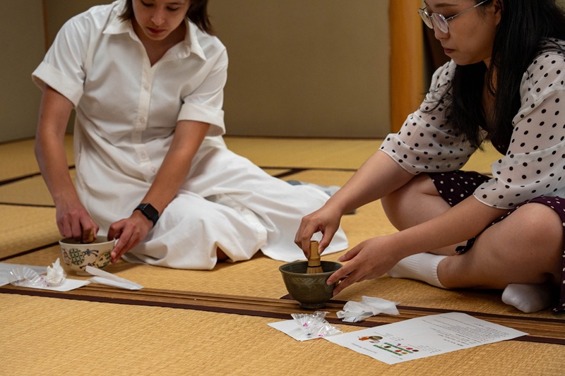 Two people in a Japanese tea ceremony, practicing how to stir matcha in a traditional tatami room.