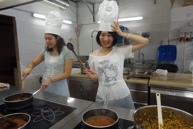 Two CET Siena students wearing a plastic apron and hat while in a commercial kitchen setting, participating in a cooking workshop.