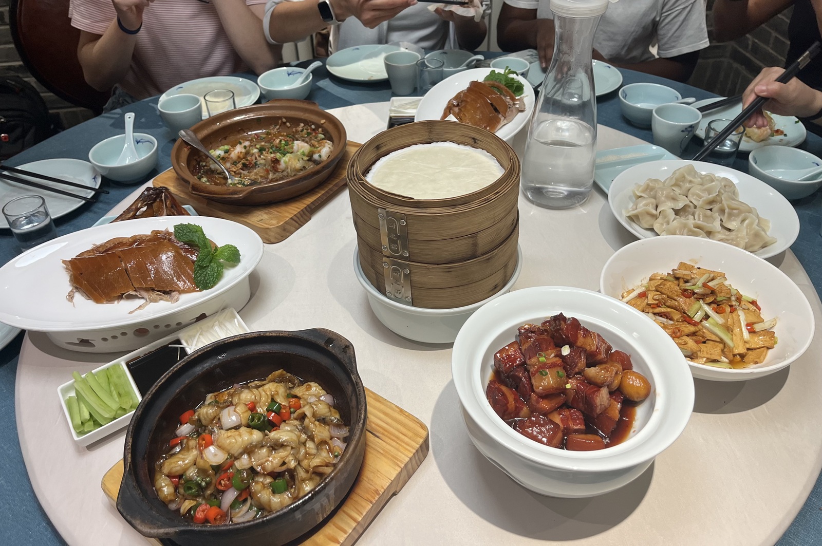 A table with a variety of Chinese dishes to eat from braised pork belly, steamed seafood stew, Peking Duck, dumplings, and shrimp with rice noodles in a clay pot.