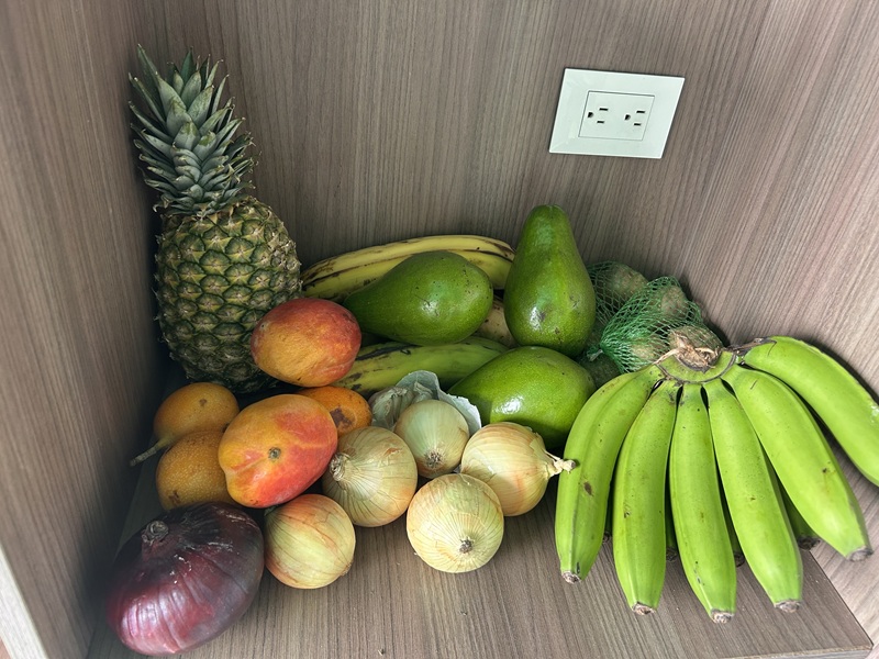 A bunch of fruits and vegetables in the corner like onions, bananas, pineapple, and avacados,