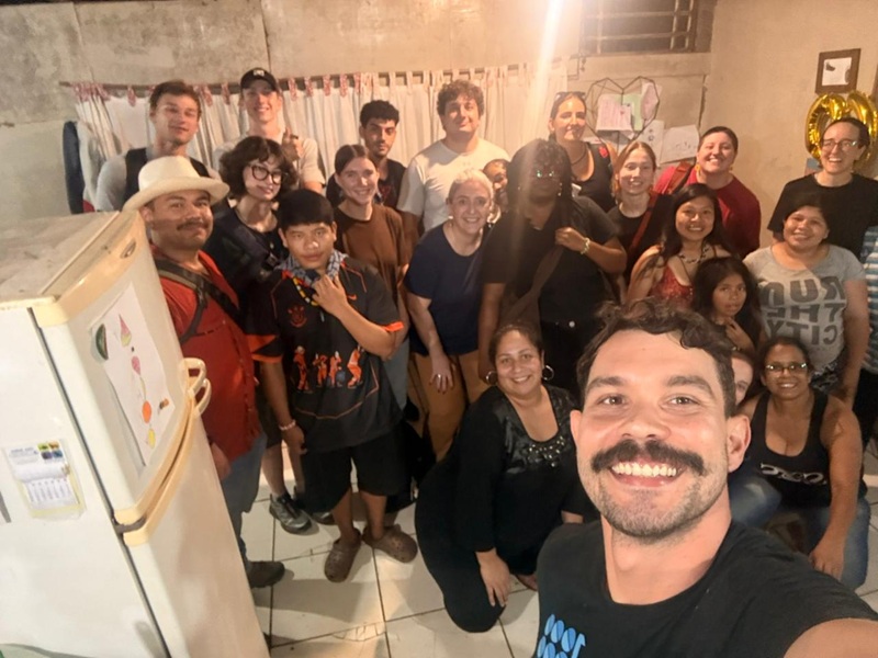 A large group selfie of community members and visitors smiling together inside a home.