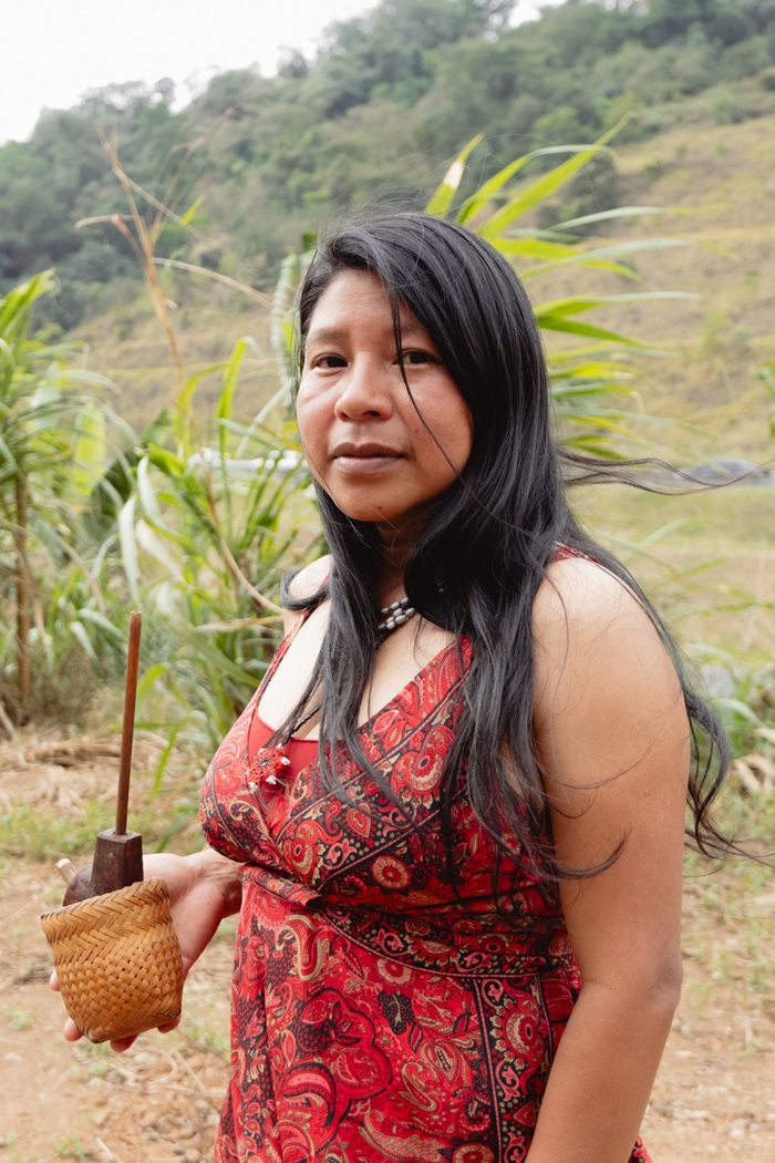 An indigenous woman in a red patterned dress holding a traditional woven pipe holder.