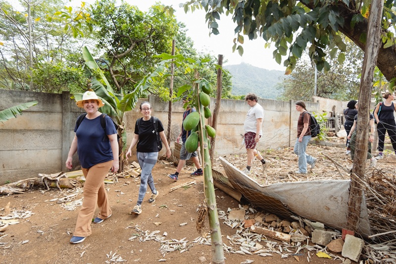 Students walking along a dirt path in a village, passing a small papaya tree with large green fruit.