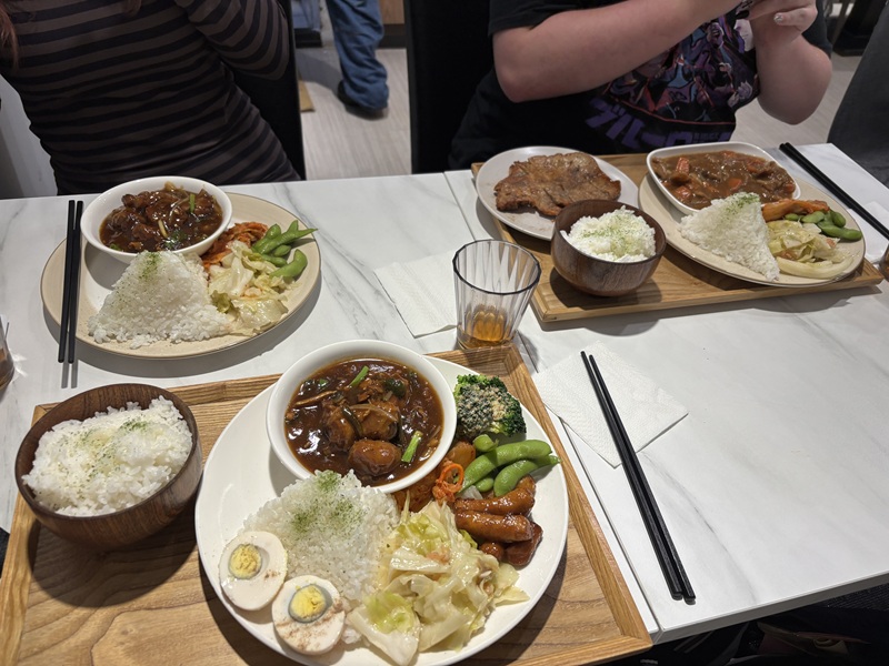 A set meal featuring a savory braised or stewed meat dish (such as braised beef or pork) served with molded white rice topped with green seasoning, halved braised eggs, cabbage, edamame, and side items on a wooden tray